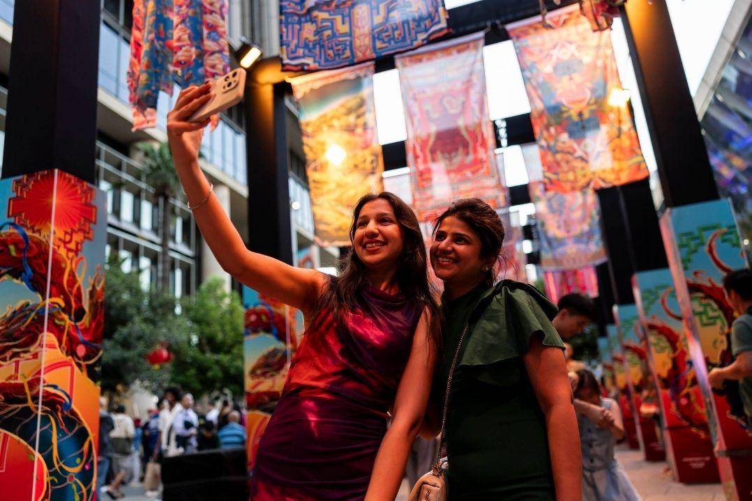 Two people smile while taking a selfie in a lively, colorful street. Vibrant banners and murals create a festive, joyful atmosphere around them.