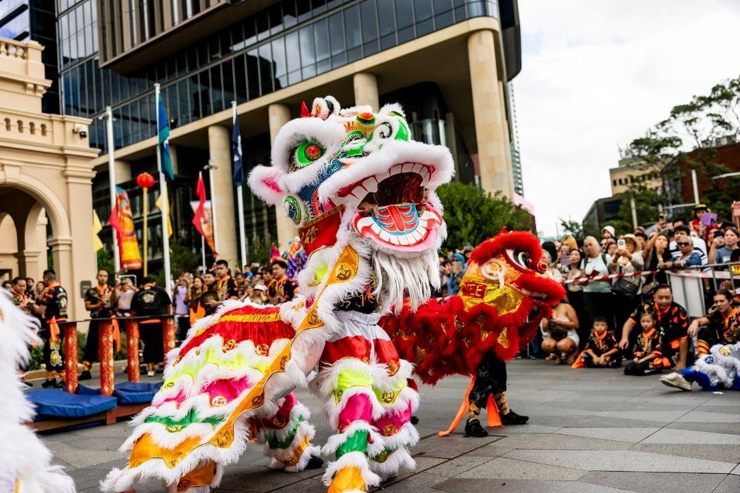 A vibrant street scene features a colorful lion dance with performers in a festive costume. A crowd watches joyfully, capturing a lively cultural celebration.