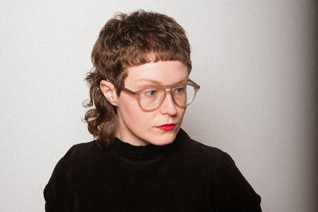 Person with short, curly hair and glasses, wearing a black turtleneck, looking to the side. The expression is thoughtful, set against a neutral background.