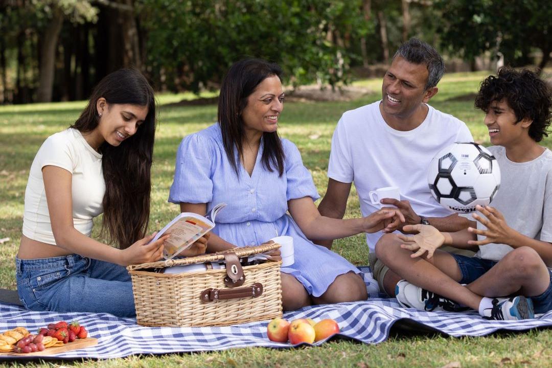 A family of four enjoys a picnic on a blue checkered blanket in a park, surrounded by trees. They are smiling, sharing food from a basket, creating a cheerful atmosphere.