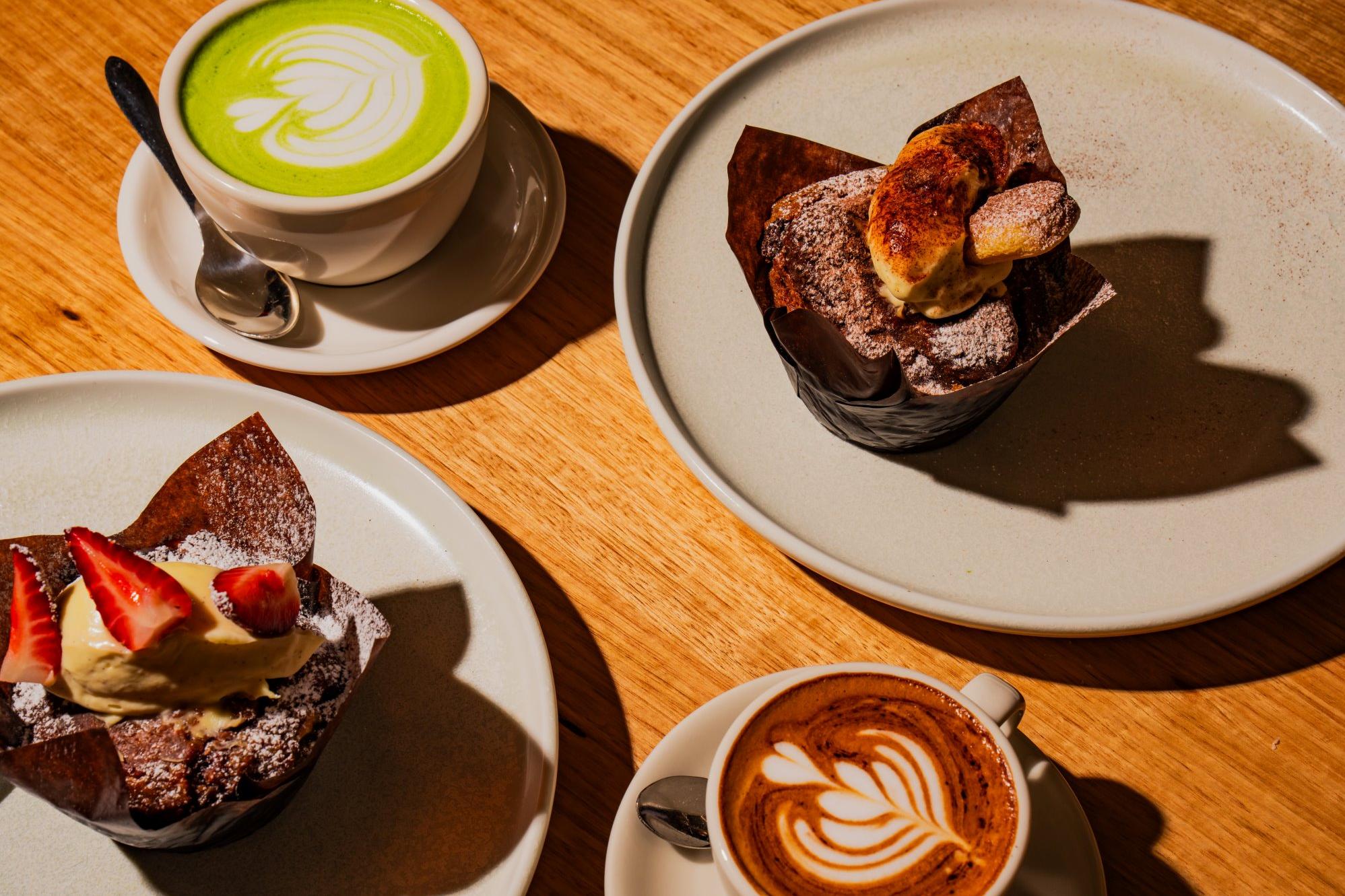 A wooden table with two muffins on plates and two lattes in cups. The lattes have intricate foam art, and one is green, indicating matcha. Warm, inviting atmosphere.