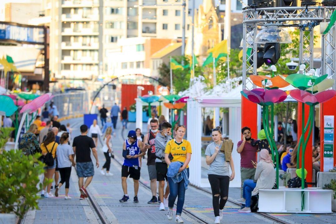 A vibrant outdoor market scene with people strolling along stalls showcasing colourful art and crafts. The atmosphere is lively and cheerful, set against urban buildings.