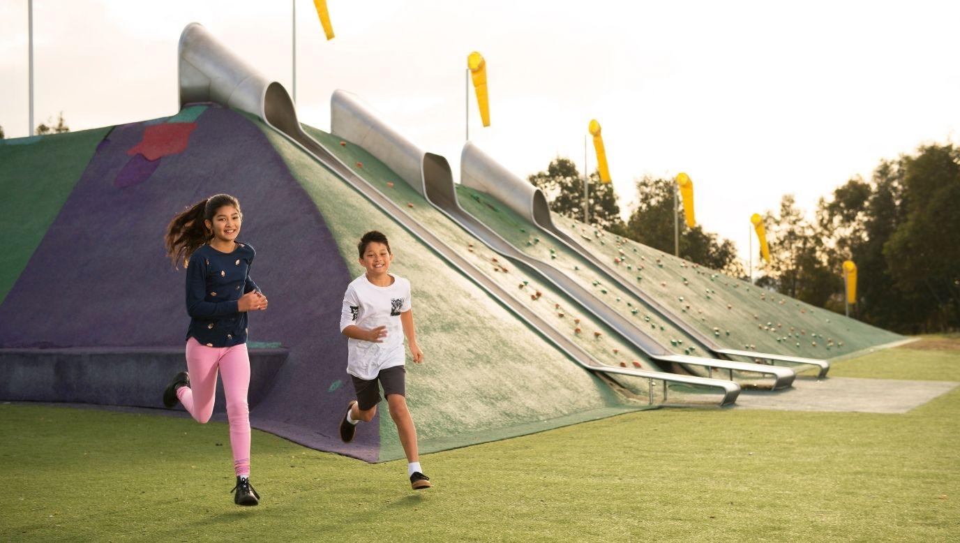 Two children joyfully race on grass near colourful slides on a clear day. The scene feels vibrant and playful, with flags waving in the background.