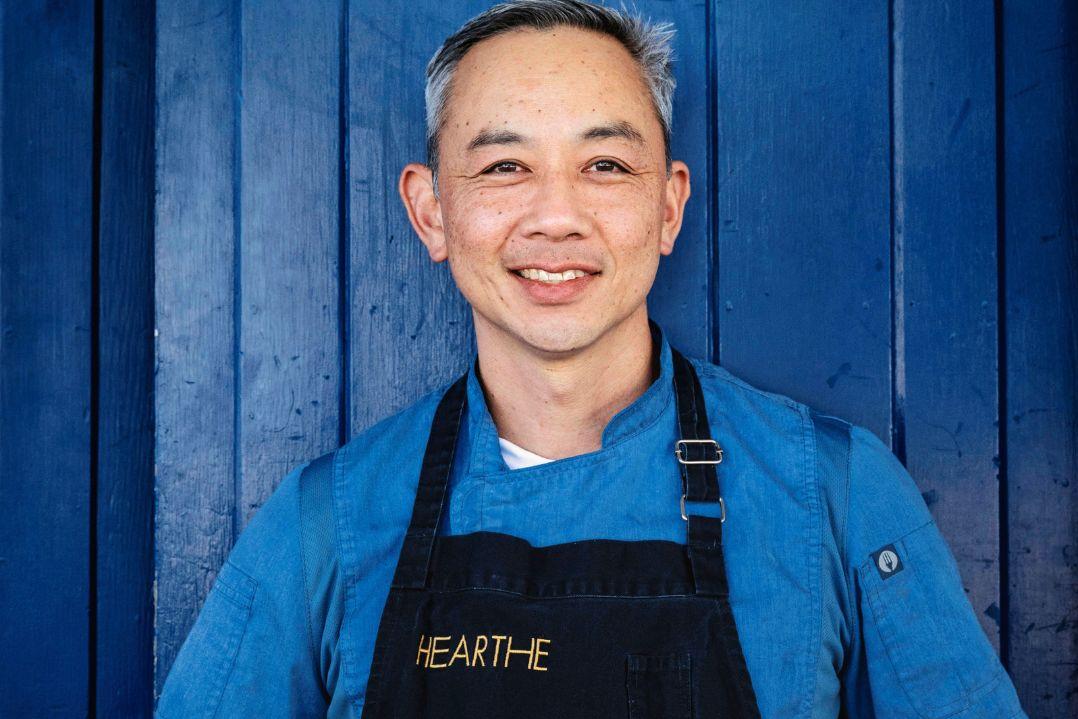 Smiling person wearing a blue shirt and black apron stands against a blue wooden background. The mood is friendly and welcoming.
