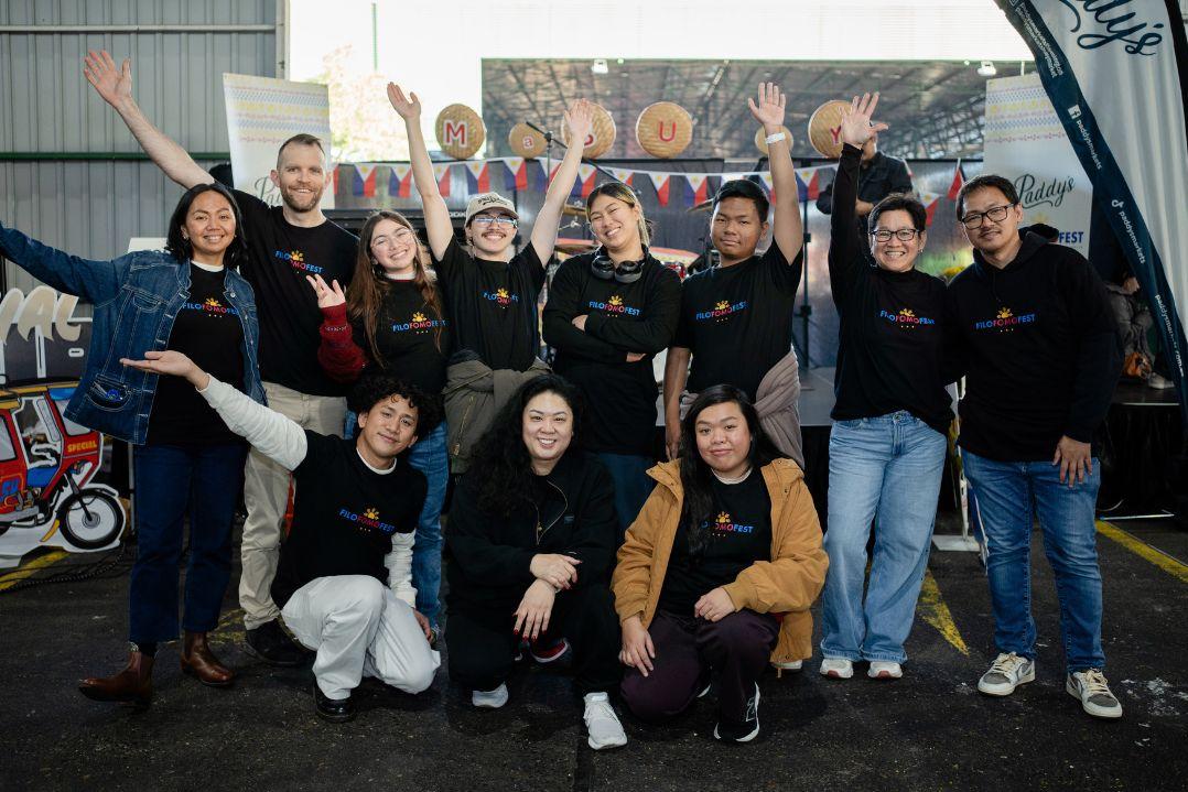 A joyful group of twelve people stand together outdoors in casual attire, smiling and raising their arms. An industrial backdrop adds to the lively atmosphere.