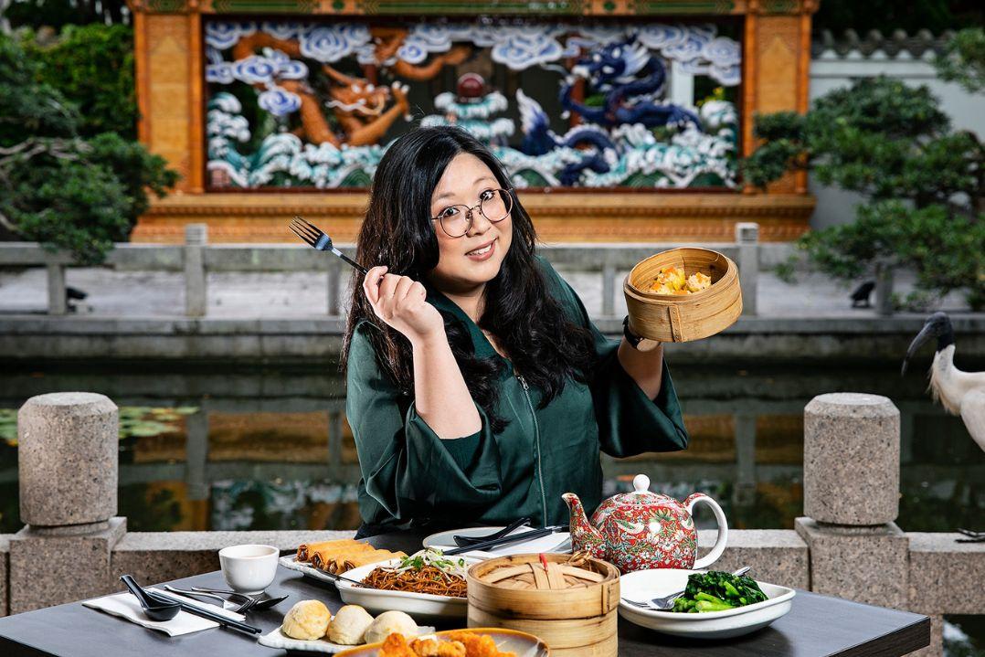 A woman with long black hair sits at an outdoor table enjoying a meal, holding a basket of oranges. Behind her is a decorative mural and greenery.