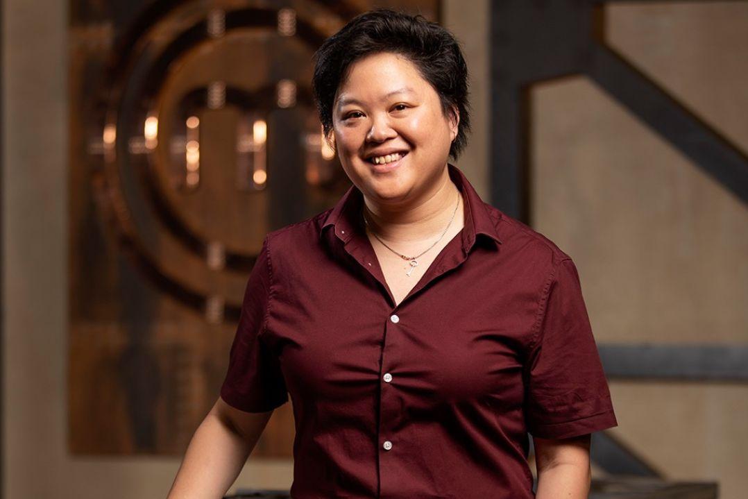 Smiling person with short hair in a burgundy shirt stands indoors, warmly lit. Background shows blurred chandelier and geometric decor, conveying a cosy ambiance.