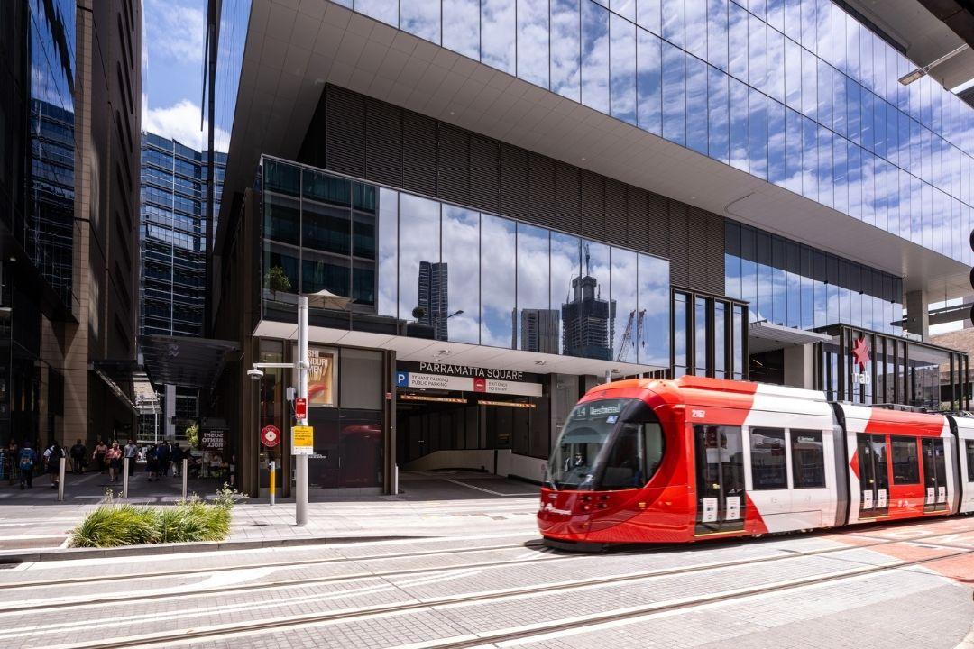 A red and white tram travels through a modern cityscape, with reflective glass buildings and a blue sky overhead, creating a dynamic urban scene.
