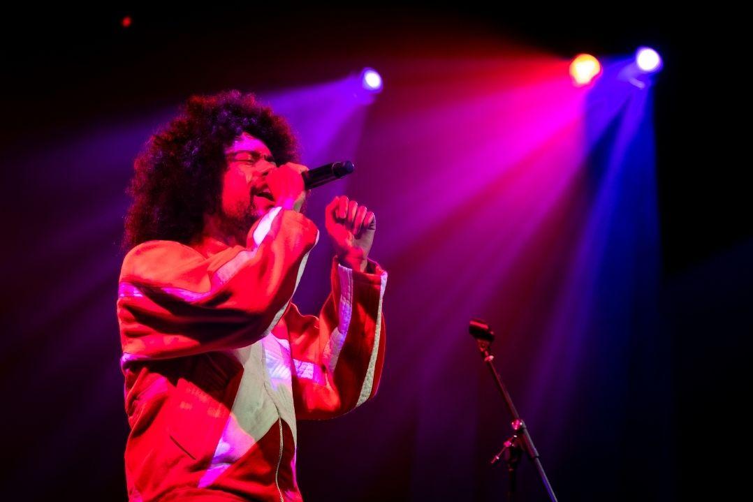 A person with curly hair sings passionately into a microphone on stage, bathed in vibrant red and purple lights, conveying energy and emotion.