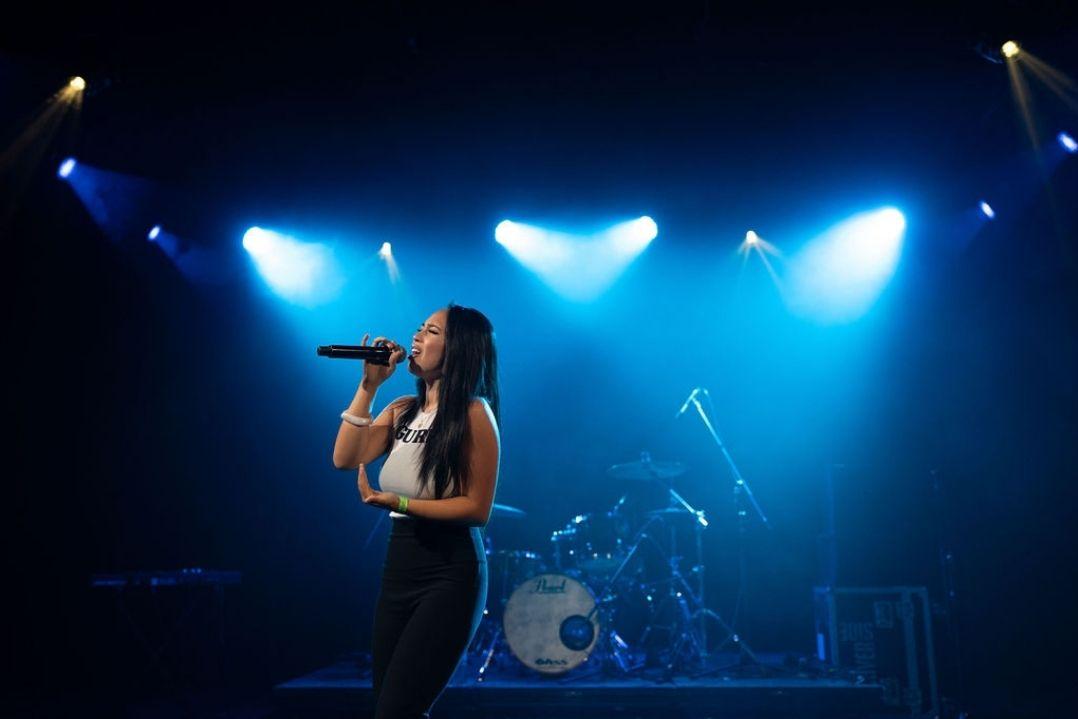 Singer passionately performing on stage under blue spotlights, holding a microphone. Drummer visible in the background, creating a vibrant concert atmosphere.