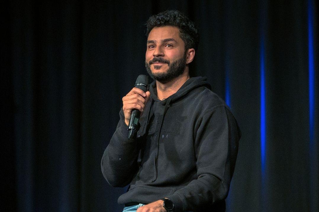 A man in a black hoodie holds a microphone on a dimly lit stage with a dark curtain backdrop, conveying a relaxed and engaging tone.