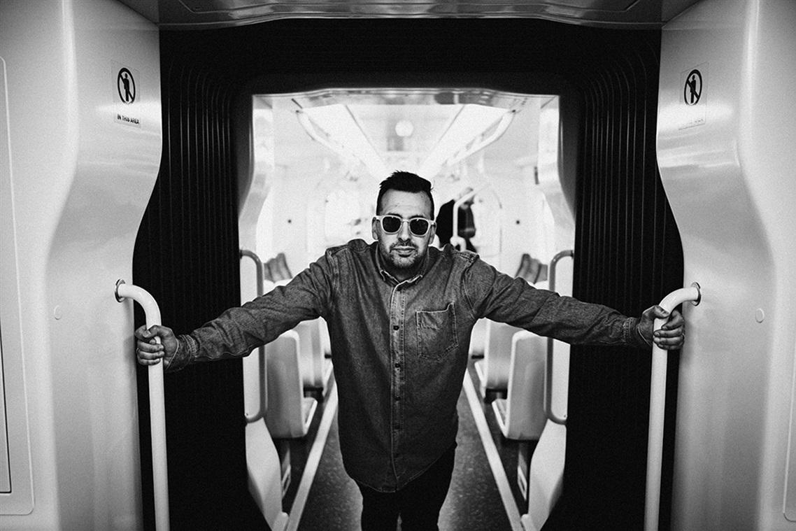 Black and white image of a person wearing sunglasses and a jacket, leaning casually with arms outstretched on a narrow walkway, conveying confidence.