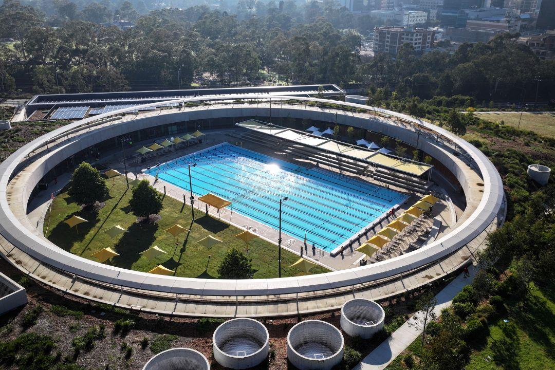 Futuristic circular building complex surrounds a large outdoor pool, with trees and pathways. Sunlight casts patterns on the sleek architecture.
