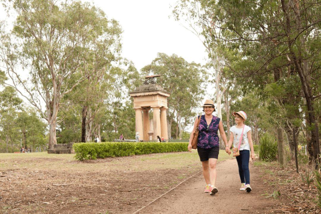 A woman and child walk on a dirt path through a wooded area. Trees surround them, and a small brick structure is visible in the background.