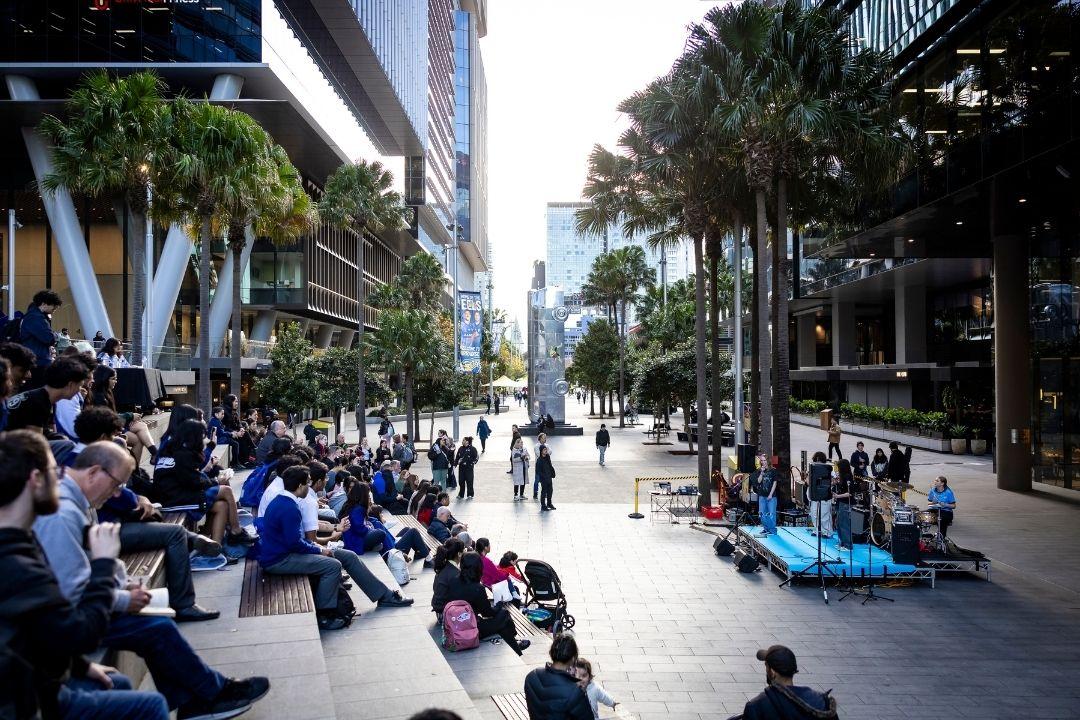A bustling urban plaza with people sitting on steps and benches beneath tall buildings and palm trees, creating a lively, relaxed atmosphere.