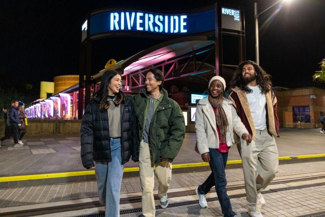 Four people stand happily under a neon-lit "Riverside" sign at night, with colorful storefronts in the background, conveying a fun, lively atmosphere.