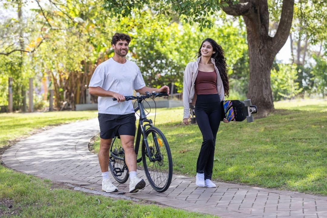 A young man, wearing a white shirt and shorts, walks a bicycle along a sunny park path. A woman with long hair follows, smiling. Both appear relaxed.