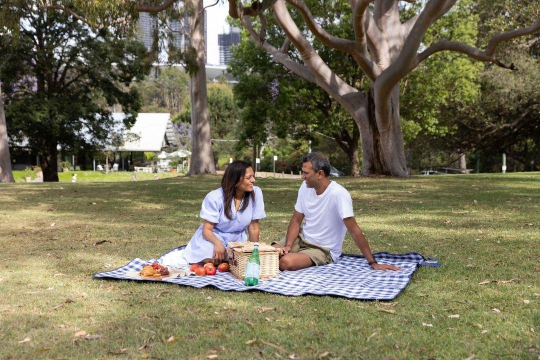 A couple enjoys a picnic on a blanket in a sunny park, surrounded by tall trees. They smile and converse, creating a warm and relaxed atmosphere.
