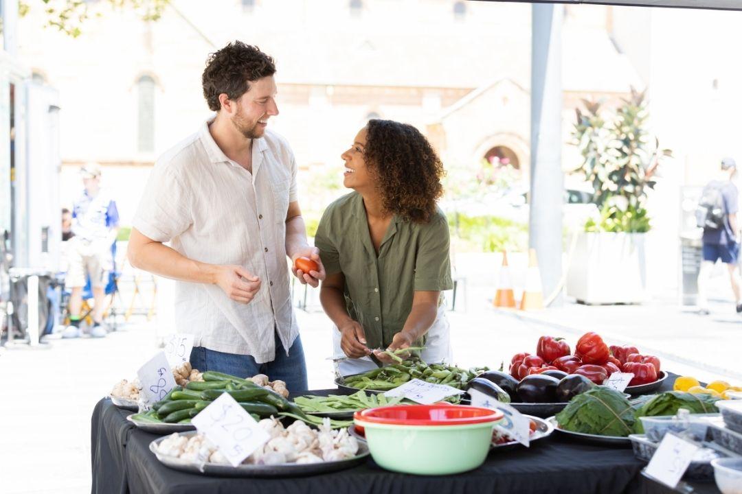 A man and woman smile and talk at a market stall with fresh vegetables, including tomatoes and leafy greens. The setting is bright and casual.