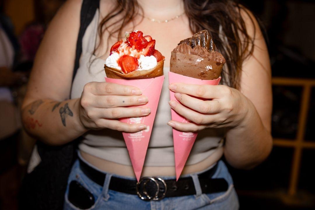A woman holds two ice cream cones with pink wrappers: one with chocolate ice cream, the other with vanilla, strawberries, and cream. Casual, indulgent mood.