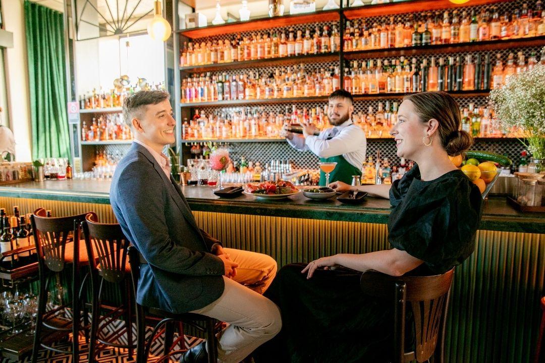 Two people sit at a rustic bar with wooden shelves filled with bottles. A bartender stands behind, smiling. The setting is cozy and inviting.
