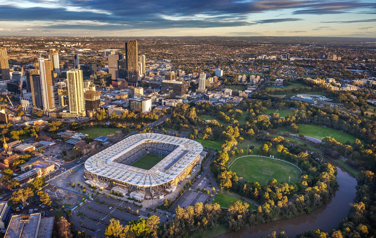 Aerial view of a cityscape at sunset, featuring a large modern stadium in the foreground, surrounded by trees and high-rise buildings in the background.