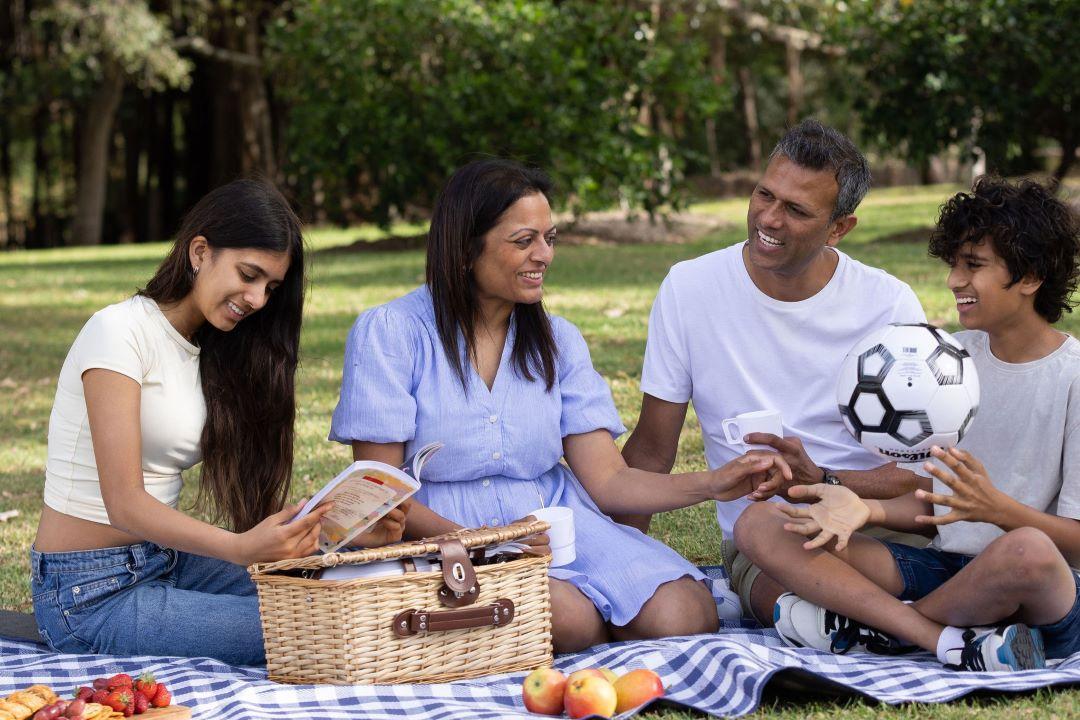 A family of four enjoys a picnic on a grassy lawn. They sit on a checkered blanket with a basket of food, surrounded by trees, smiling and chatting.
