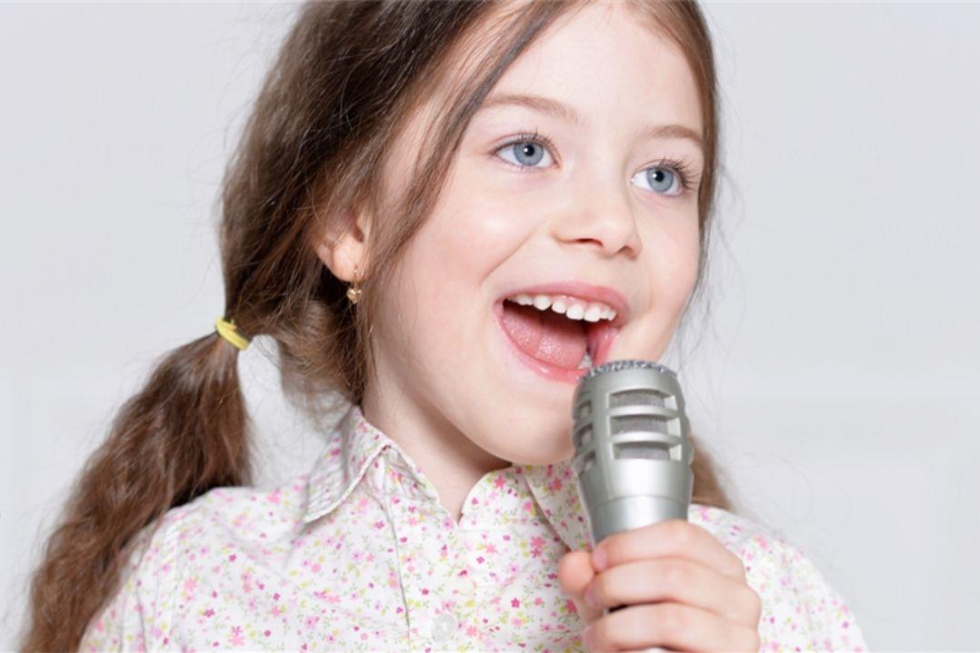 A young girl with brown hair in pigtails sings joyfully into a microphone. She wears a light pink top, expressing enthusiasm and happiness.