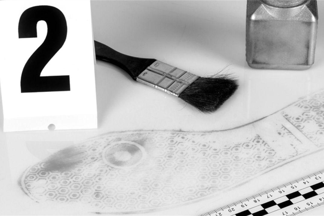 Close-up of a crime scene with a large number "2" marker, a paintbrush, transparent liquid bottle, and shattered glass on a textured surface.