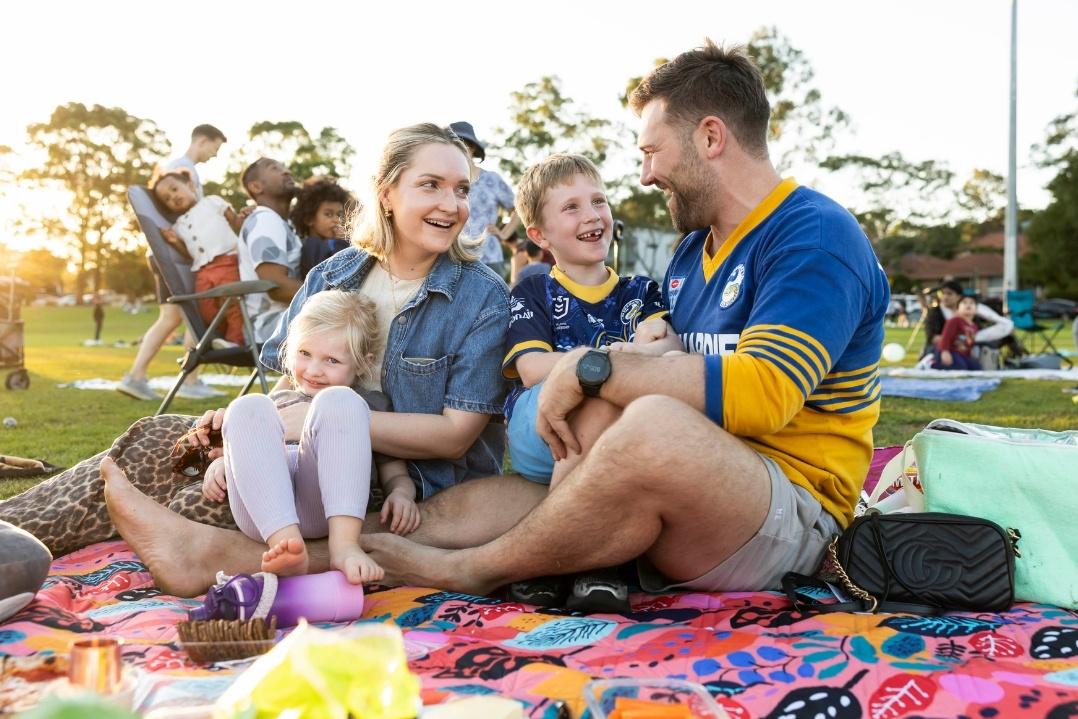 A family of four enjoys a picnic on a checkered blanket in a sunny park. They laugh and smile, surrounded by trees and other people in the background.