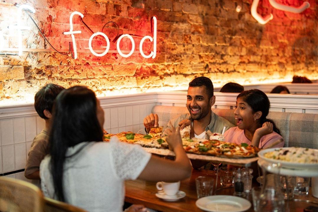 Four people sit around a restaurant table, laughing and enjoying a meal. Neon "food" sign glows on brick wall, creating a warm, cheerful atmosphere.