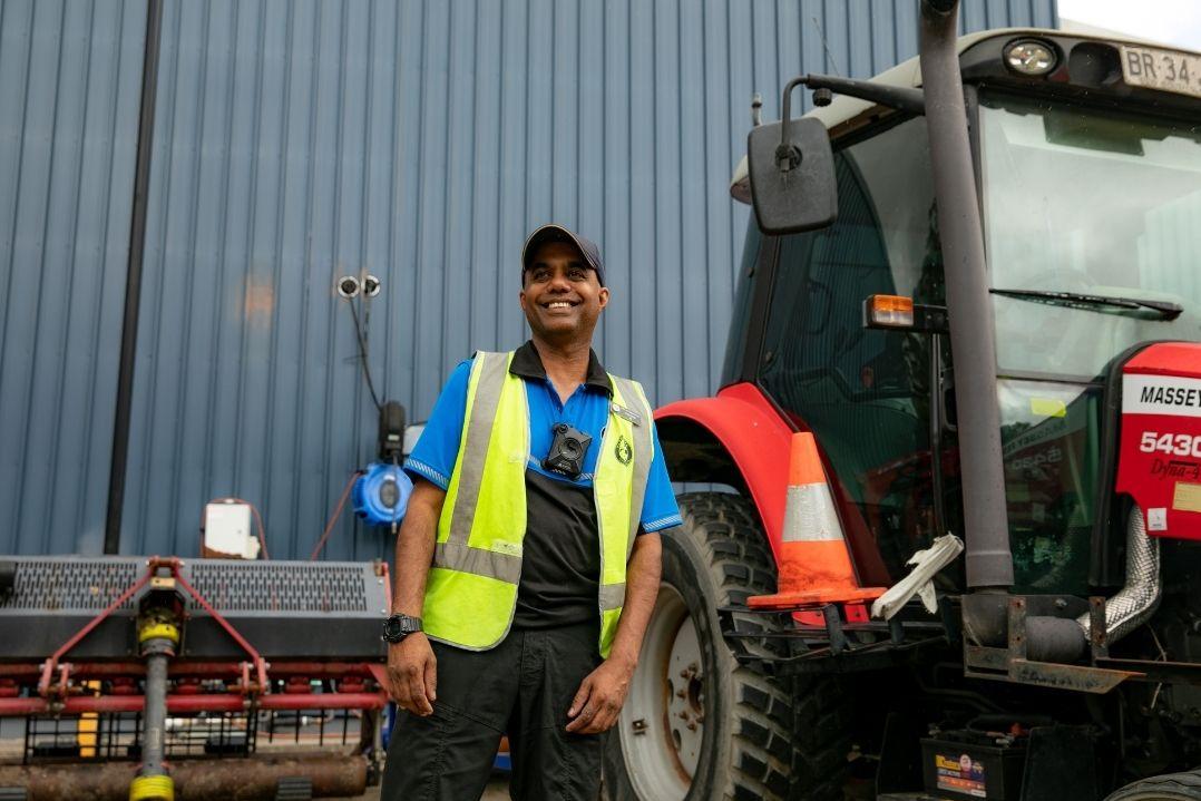 A man wearing a yellow safety vest and blue shirt stands confidently beside a red tractor in front of a large corrugated metal wall. The scene conveys a sense of professionalism and industrial work.