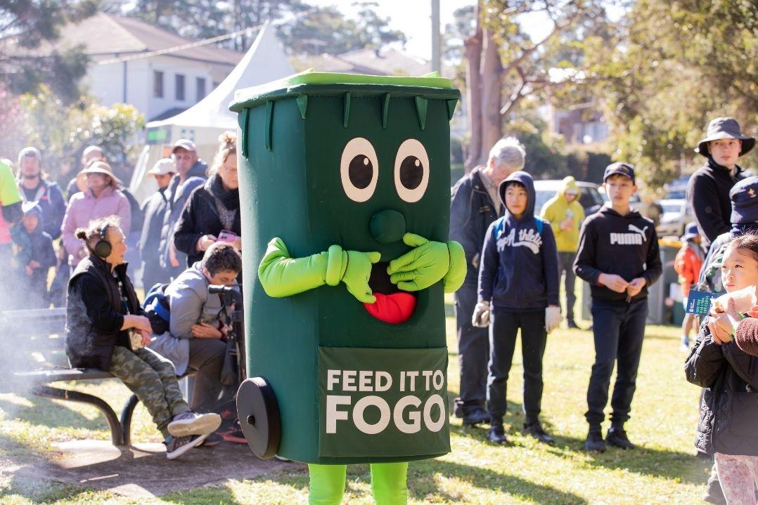 A person in a green recycle bin costume with big eyes and red lips, labeled "Feed Me To FOGO," interacts with onlookers at an outdoor event, creating a playful atmosphere.