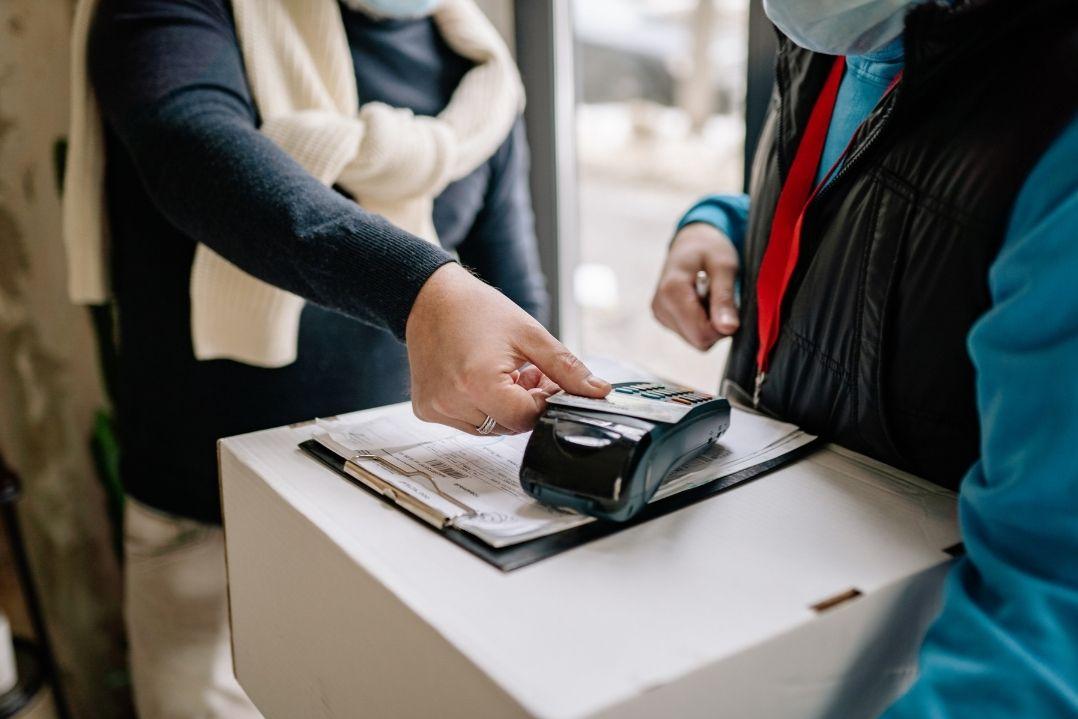 A person uses a contactless payment card on a terminal held by another individual outdoors. Both are dressed warmly, suggesting cold weather.