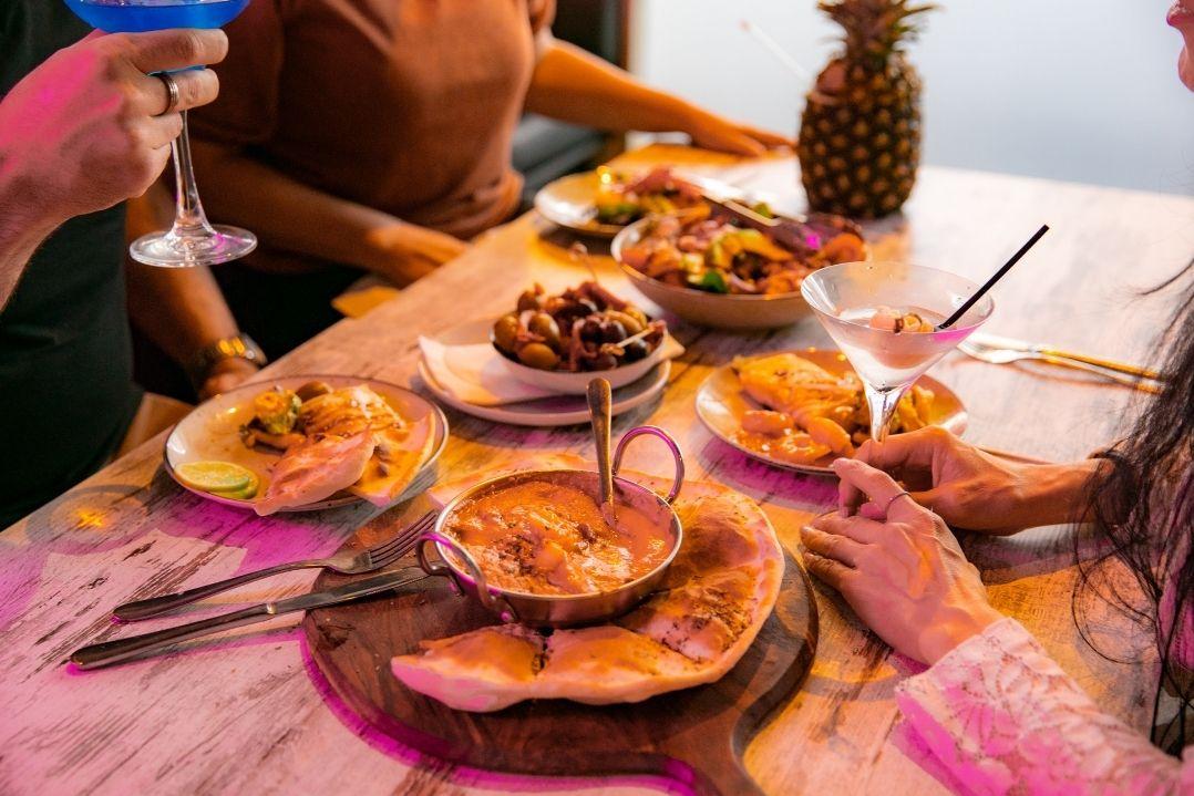 A cozy dinner table with pink lighting, featuring pizzas, fries, and a chocolate dessert. Diners toast with wine glasses, creating a warm, celebratory atmosphere.