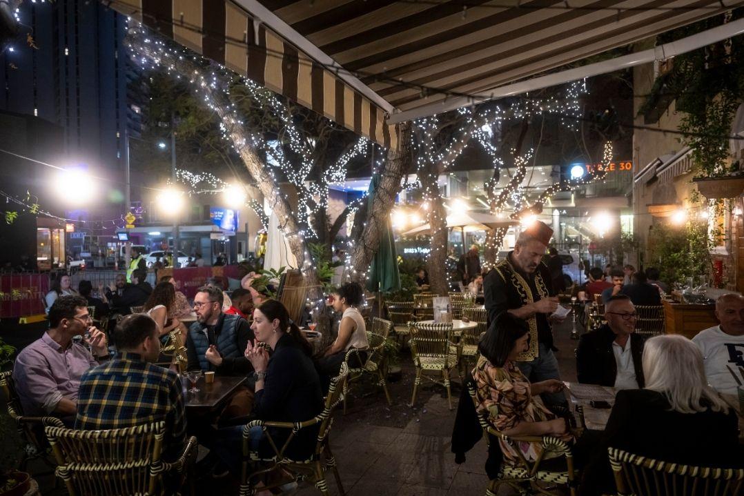 Outdoor restaurant bustling with people dining under string lights at night. The atmosphere is lively and warm, with wooden beams and foliage.
