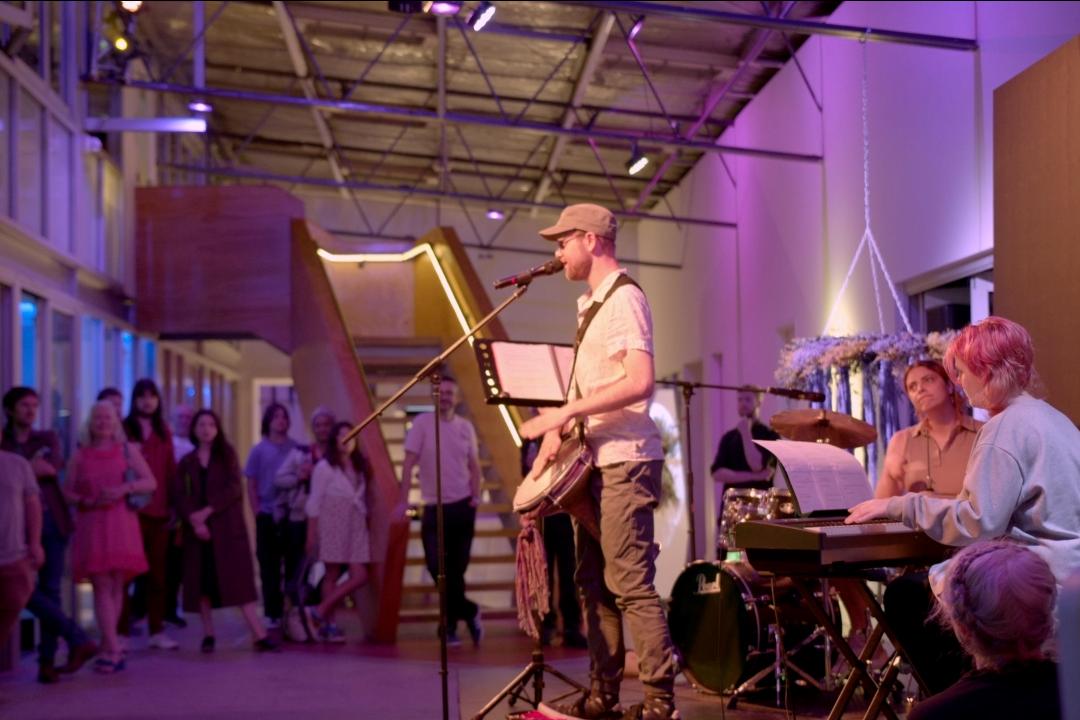 A jazz band performs in a warmly lit venue. The saxophonist stands at the center, with a pianist and drummer nearby. An engaged audience listens.