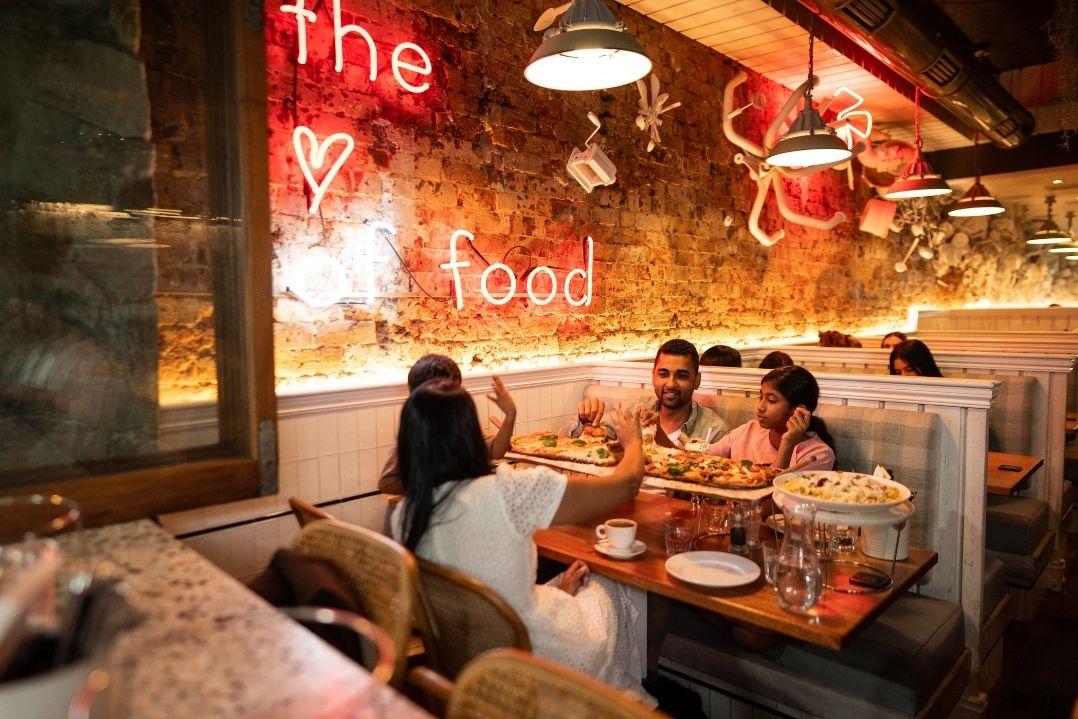 A family of four smiles and poses for a selfie in a restaurant booth. Warm, cozy lighting and a neon "Food" sign create a cheerful atmosphere.
