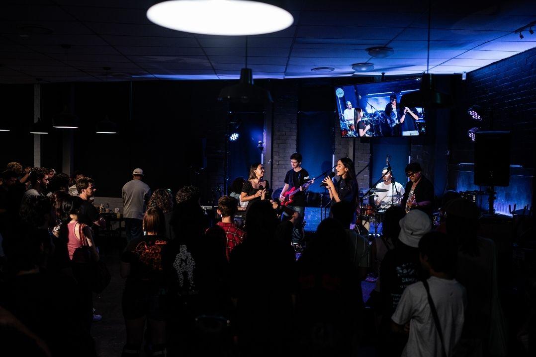 A band performs on a dimly lit stage with blue lights, surrounded by an audience. The atmosphere is vibrant and energetic, with people enjoying the music.