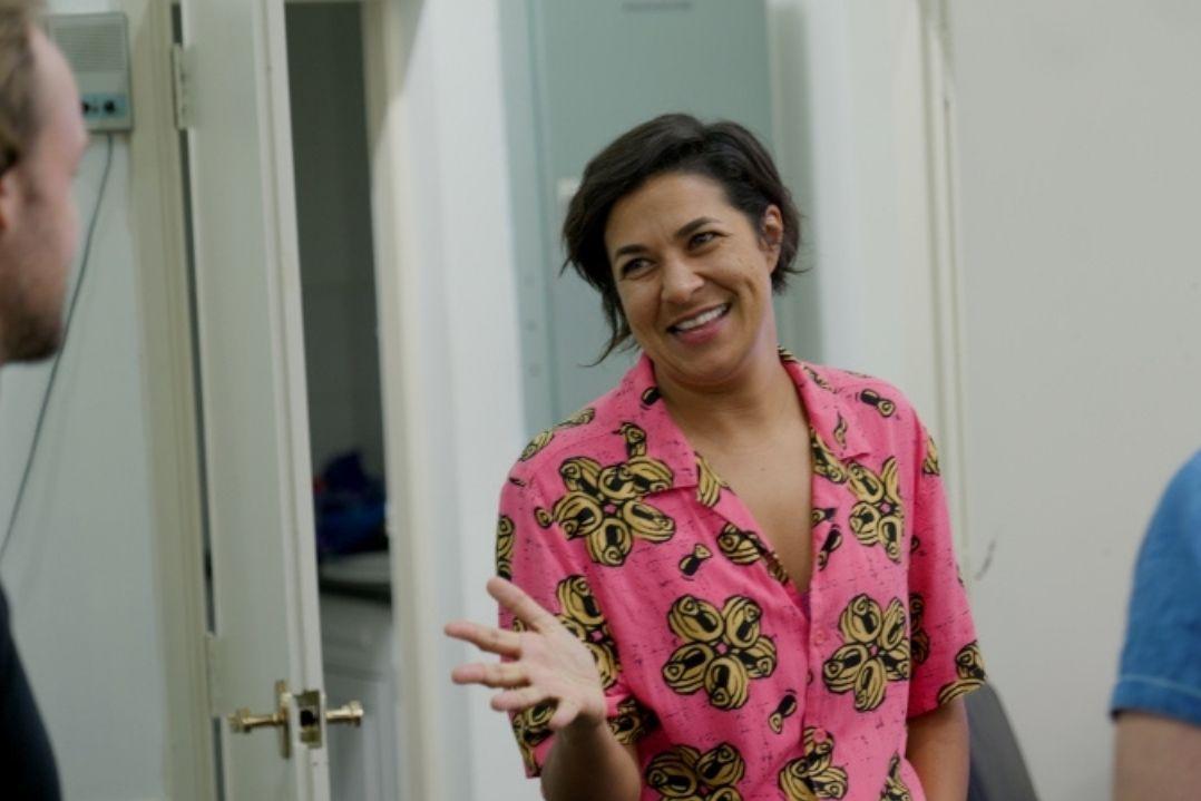Woman in a vibrant pink, floral top stands indoors, smiling warmly at two people partially shown. The atmosphere is friendly and inviting.