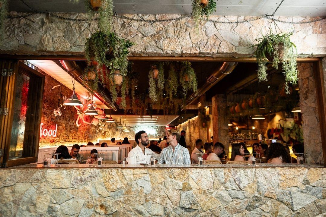 A bustling, warmly lit restaurant with diners seated and green plants hanging from the ceiling, creating a lively and inviting atmosphere.