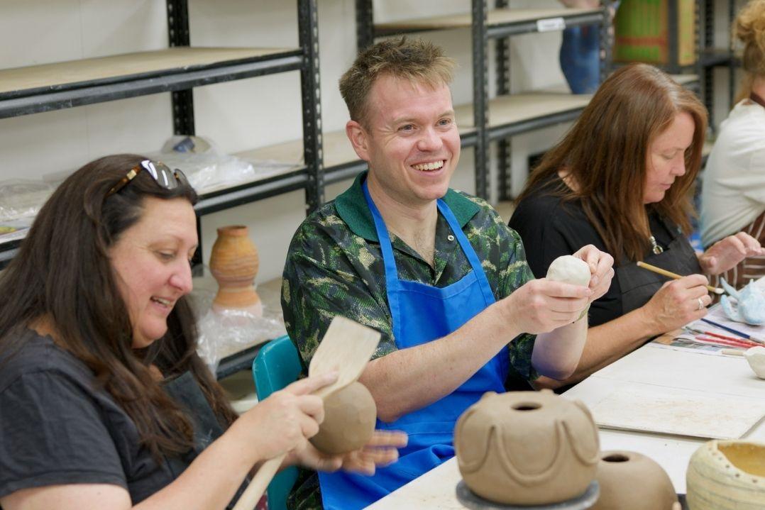 Three people smiling and shaping clay in a pottery class. They sit at a table with pottery tools and shelves of pottery in the background, enjoying the creative process.