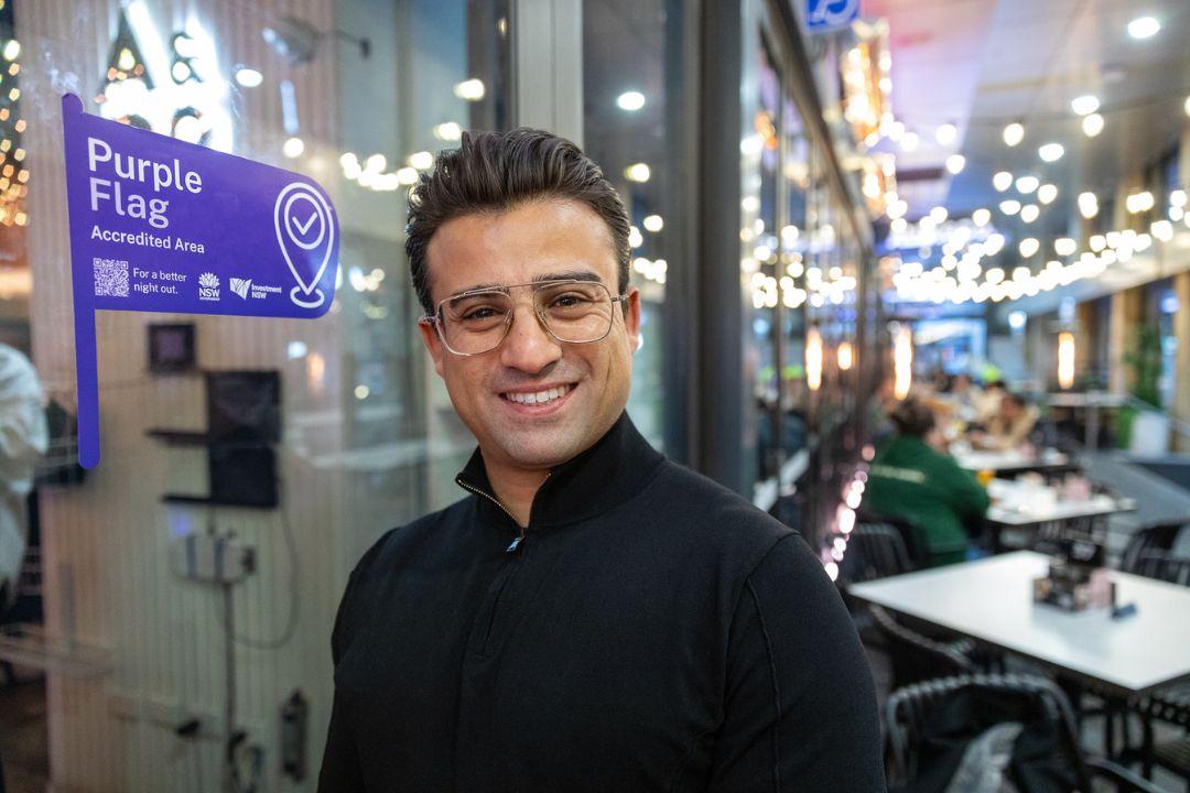 Smiling person in a black shirt stands in a modern, brightly-lit office with glass walls, open desks, and a few people working in the background.