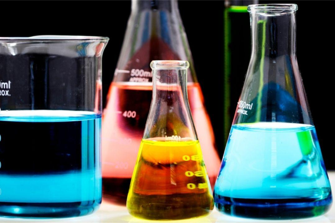 Three laboratory flasks, each with colorful liquids: blue in a beaker, orange in an Erlenmeyer flask, and red in a conical flask, on a dark background.