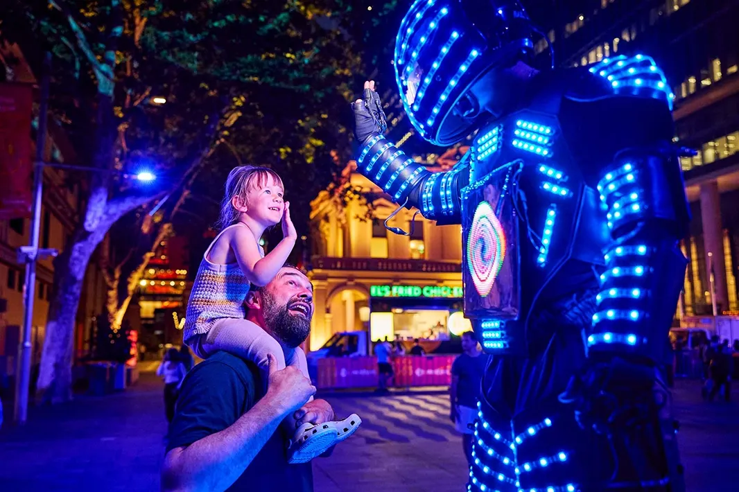 A man carries a joyful girl on his shoulders, both smiling at a performer in a glowing, futuristic costume.