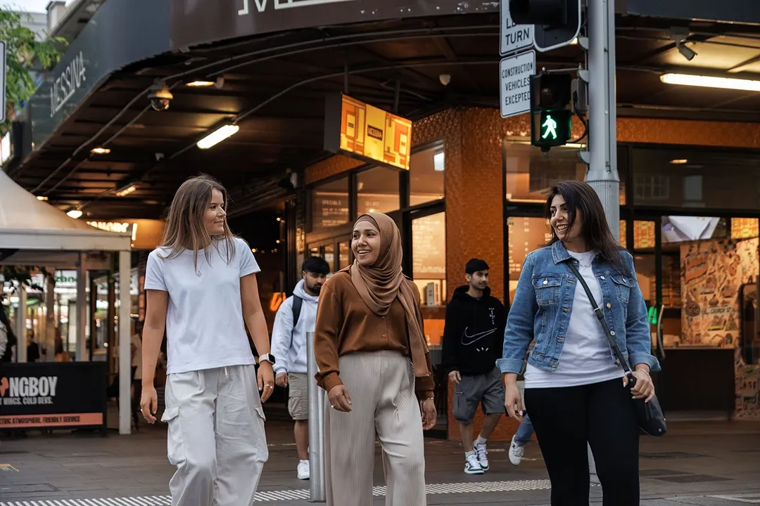 Three women smiling and chatting as they cross a city street at dusk. The scene is lively, with bustling shops and a warm atmosphere.