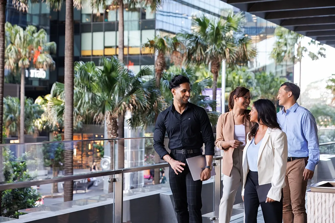 A group of four professionals, three women and a man, are walking and conversing on a city balcony with palm trees and modern glass buildings behind them.