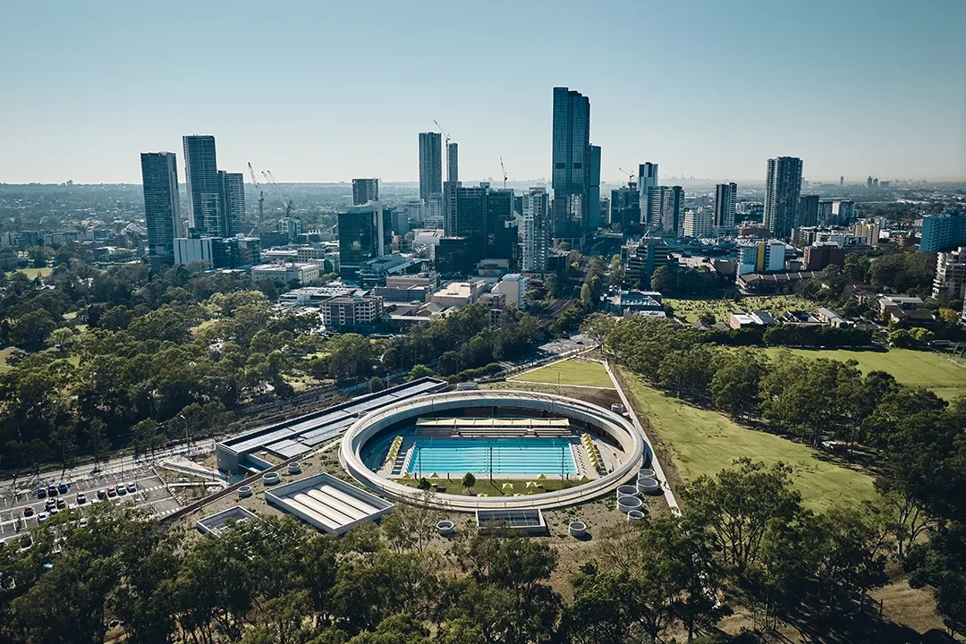 Aerial view of a circular outdoor swimming pool amid lush greenery, set against a backdrop of tall city skyscrapers under a clear blue sky.