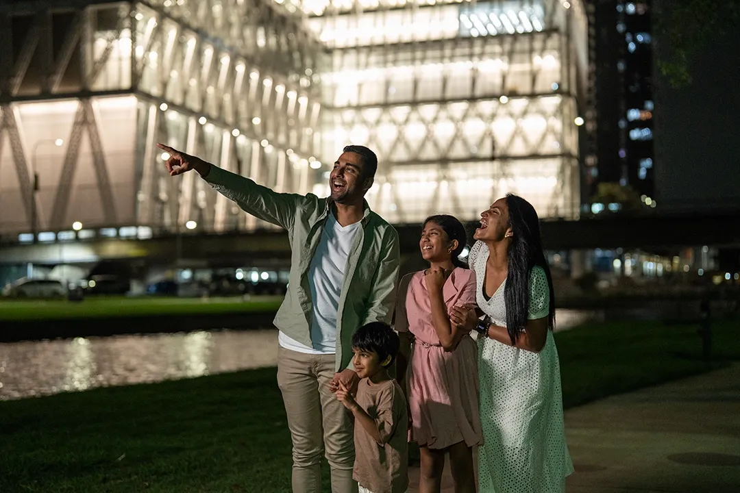 A joyful family of four stands near a lit modern building at night, with the father pointing excitedly.