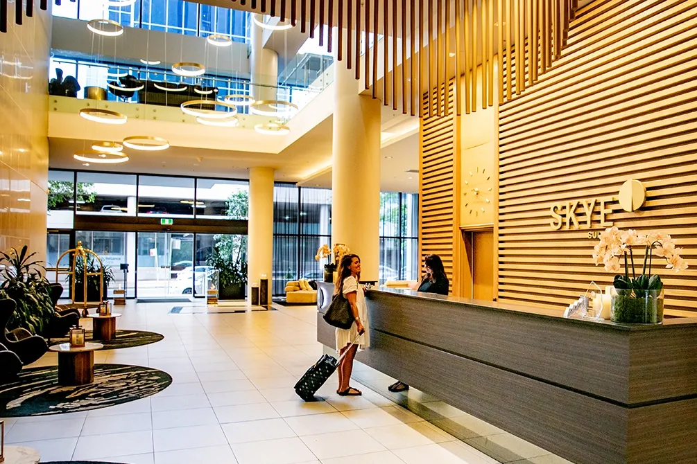 Hotel lobby with modern decor. A receptionist assists a guest at the wooden front desk. Elegant lighting, plants, and sofas create a welcoming ambiance.